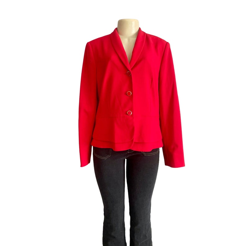 May include: A bright red blazer featuring a shawl collar, long sleeves, and three gold-toned buttons down the front. The jacket has a layered hemline detail and is displayed on a mannequin paired with dark charcoal gray denim jeans.