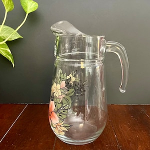 Puede incluir: Una jarra de vidrio transparente con un dise&ntilde;o floral con flores rosas, amarillas y verdes. La jarra tiene un asa y una boca ancha.