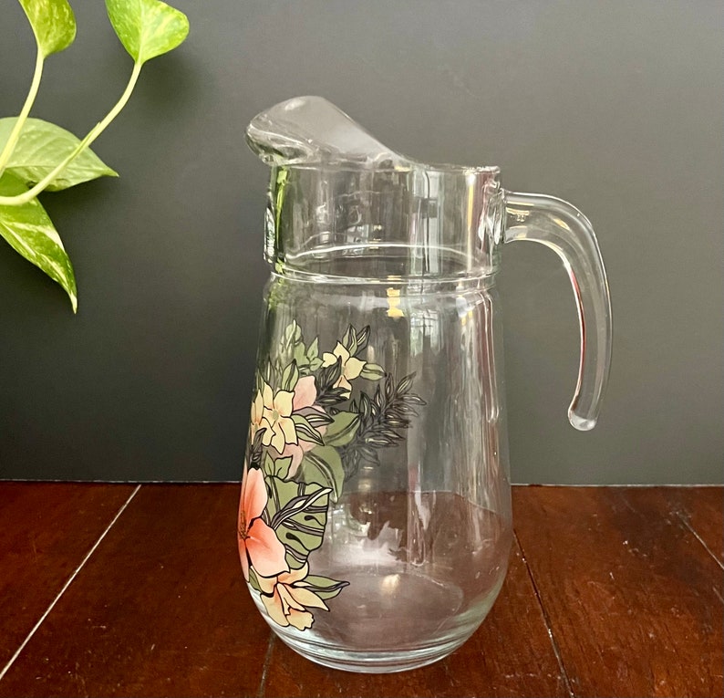 Puede incluir: Una jarra de vidrio transparente con un dise&ntilde;o floral con flores rosas, amarillas y verdes. La jarra tiene un asa y una boca ancha.