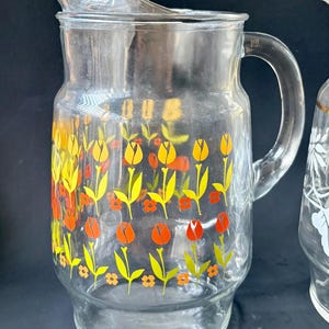 May include: A clear glass pitcher with a floral design. The pitcher has a wide mouth and a handle. The floral design features yellow, orange, and green tulips.