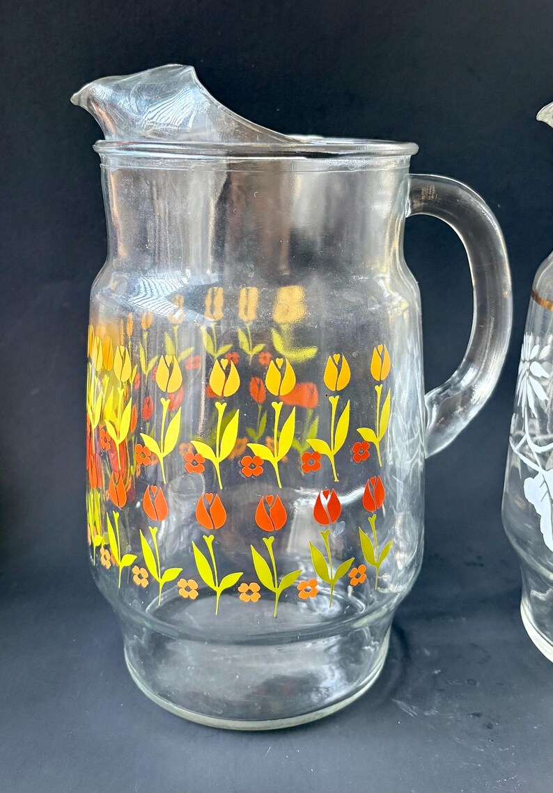 May include: A clear glass pitcher with a floral design. The pitcher has a wide mouth and a handle. The floral design features yellow, orange, and green tulips.