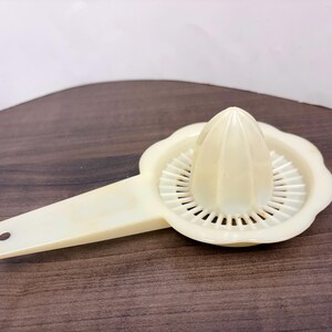 May include: A vintage, off-white plastic citrus juicer. The juicer has a reamer, a strainer, and a handle with a hole for hanging. The juicer is designed to extract juice from citrus fruits like lemons and limes.