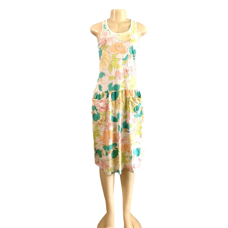 May include: A white sleeveless dress with a floral print in shades of pink, green, and yellow. The dress has a gathered waist and two large pockets on the front.