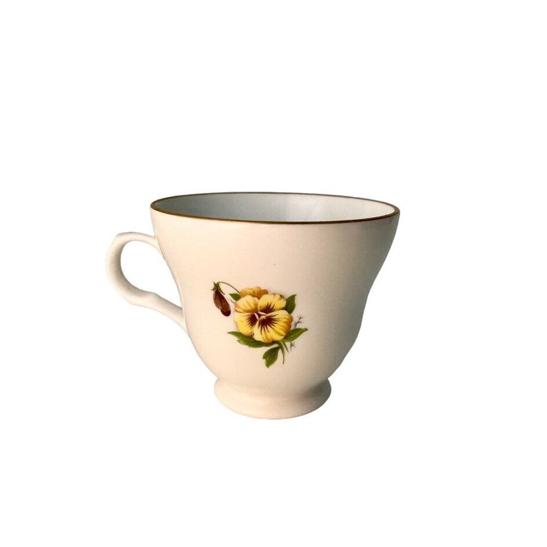 May include: Cream-coloured teacup with a yellow and brown pansy floral design. The cup has a gold rim and a small handle. The teacup is on a small pedestal base.