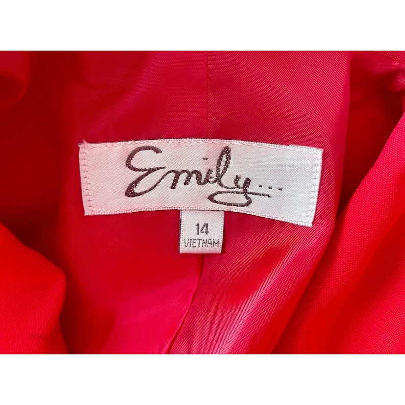 May include: A bright red garment interior featuring a white rectangular clothing label with the text Emily in dark brown script. A smaller white tag attached below displays the number 14 and the word VIETNAM in black capital letters.