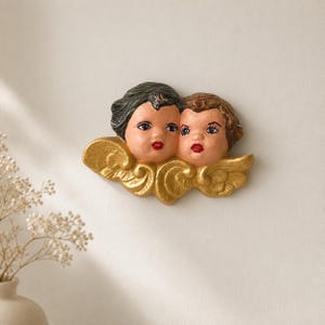 May include: A wall-mounted decorative plaque featuring two cherub faces with painted features, including dark hair, light skin, and red lips, resting atop a pair of sculpted, gold-colored wings with detailed feather textures and swirling patterns.