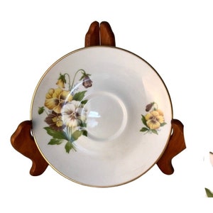 May include: White saucer with a gold rim and a floral design featuring yellow, purple, and white pansies. The saucer is displayed on a wooden stand, showcasing the delicate floral pattern.