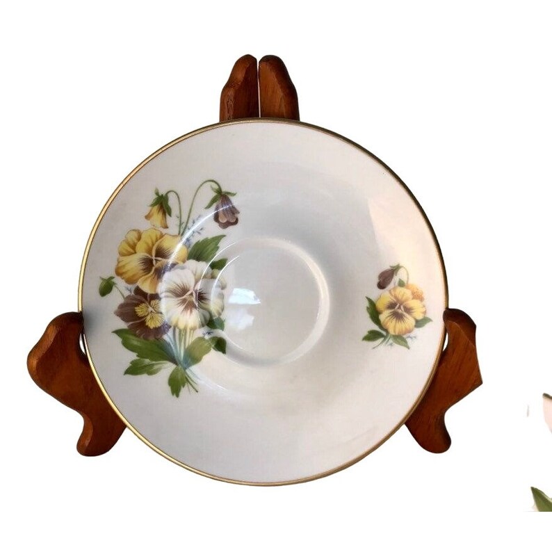 May include: White saucer with a gold rim and a floral design featuring yellow, purple, and white pansies. The saucer is displayed on a wooden stand, showcasing the delicate floral pattern.
