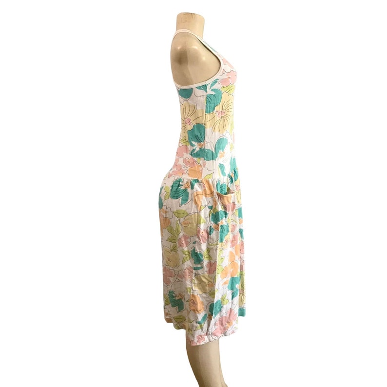 May include: A white sundress with a floral pattern in shades of pink, yellow, green, and blue. The dress has a scoop neck, a fitted bodice, and a knee-length skirt with pockets. The dress is displayed on a mannequin.