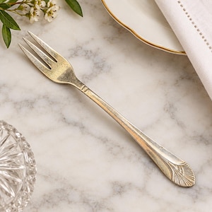 May include: A silver-toned metal fork with three tines and a tapered handle featuring a fan-shaped embossed pattern at the base. The metal surface shows a weathered, aged patina with subtle variations in color and texture along the stem.