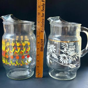 May include: Two glass pitchers with floral designs. One pitcher has a yellow, orange, and green floral design. The other pitcher has a white floral design with a gold band around the middle.