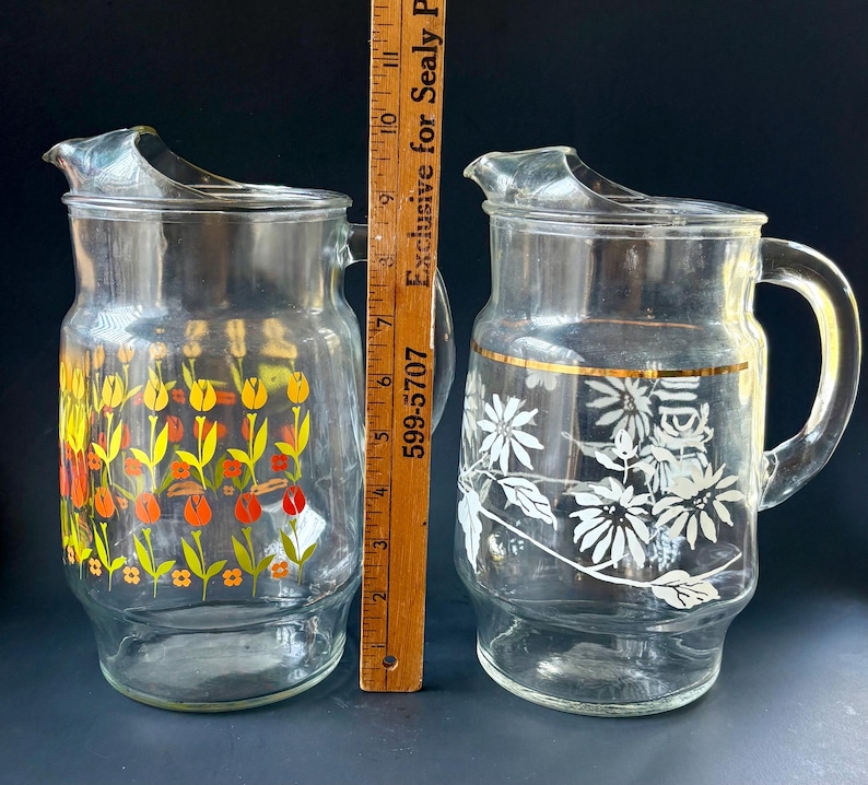 May include: Two glass pitchers with floral designs. One pitcher has a yellow, orange, and green floral design. The other pitcher has a white floral design with a gold band around the middle.