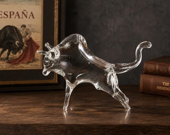 May include: A clear glass figurine of a bull in a charging stance. The sculpture features a smooth, transparent surface with defined horns, a muscular hump, and a curved tail extended upwards, capturing the animal in a dynamic, forward-leaning pose.