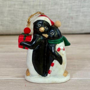 May include: A Christmas ornament featuring two penguins. One penguin wears a Santa hat and holds a red gift box. The other penguin wears a green scarf and has a candy cane. The ornament is painted in black, white, red, and gold.