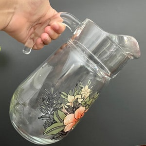 Puede incluir: Una jarra de vidrio transparente con un dise&ntilde;o floral. La jarra tiene un asa y una boca ancha. Las flores son rosas, blancas y verdes.