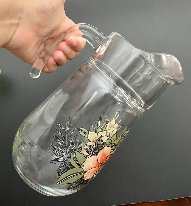 Puede incluir: Una jarra de vidrio transparente con un dise&ntilde;o floral. La jarra tiene un asa y una boca ancha. Las flores son rosas, blancas y verdes.