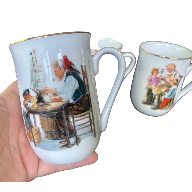 May include: Two white ceramic mugs with gold-toned rims. The larger mug features a colorful illustration of a person with a red bird on their shoulder working on a model ship at a table. The smaller mug shows a scene with two people sitting together.