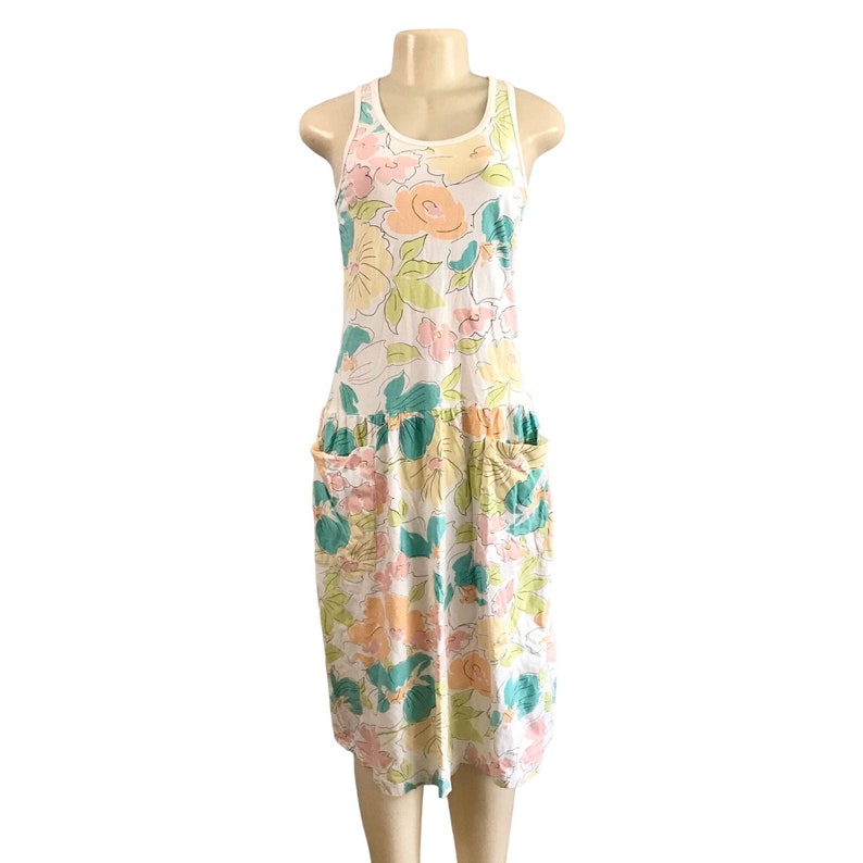 May include: A sleeveless floral dress with a white background. The dress features a scoop neck and two front pockets. The floral pattern includes pink, yellow, and teal flowers. The dress is displayed on a mannequin.