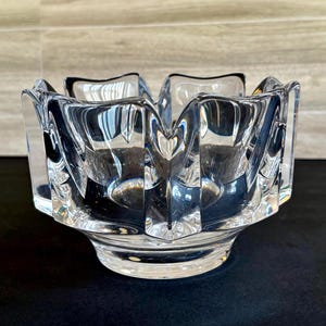 Puede incluir: Un cenicero de cristal transparente y facetado con forma de flor. El cenicero tiene cuatro secciones y una base gruesa y redondeada. El cristal refleja la luz, creando un efecto brillante. El cenicero es un artículo decorativo.