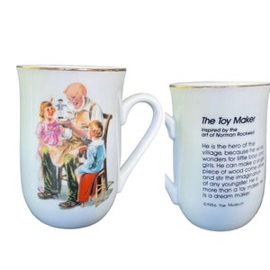 May include: A white ceramic mug with a gold rim featuring a Norman Rockwell illustration of a toy maker with two children. The reverse side displays the text: The Toy Maker inspired by the art of Norman Rockwell. He is the hero of the village.