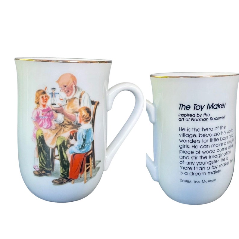 May include: A white ceramic mug with a gold rim featuring a Norman Rockwell illustration of a toy maker with two children. The reverse side displays the text: The Toy Maker inspired by the art of Norman Rockwell. He is the hero of the village.