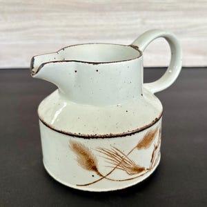 May include: A white ceramic creamer with a brown wheat design. The creamer has a rounded body and a handle.