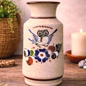 May include: A cylindrical ceramic vase with a speckled beige finish, featuring a hand-painted design of a central owl surrounded by blue flowers, green leaves, and brown fruit motifs, accented by dark brown horizontal bands at the rim and base.