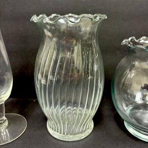 May include: Clear glass vase with vertical ribbed design and a ruffled top. The vase is cylindrical with a flared base and a slightly wider top. The glass has a slight green tint. Other glass objects are visible in the background.