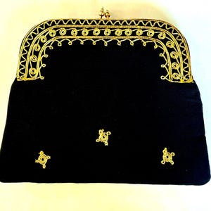May include: Black clutch with gold-colored trim and accents. The frame and clasp are gold-toned, with intricate gold thread detailing around the edges. Small gold embellishments are scattered across the front of the bag.