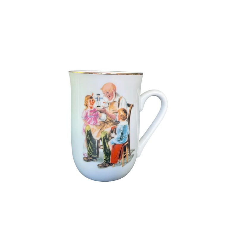 May include: A white ceramic mug with a gold-toned rim and a handle on the right side. The front features a colorful illustration of an elderly person with a white beard sitting in a chair, painting a small toy while two children watch closely.