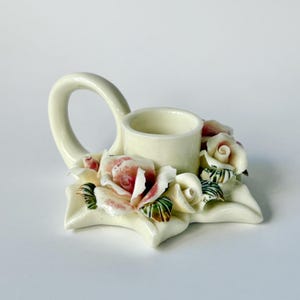 May include: Cream-coloured ceramic candle holder with a handle and a floral design. The base is shaped like a leaf, adorned with pink and white roses and green leaves. The candle holder has a circular opening.