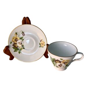 May include: A white teacup and saucer set decorated with yellow and purple pansies. The cup and saucer have gold edging. The saucer is displayed on a wooden stand.