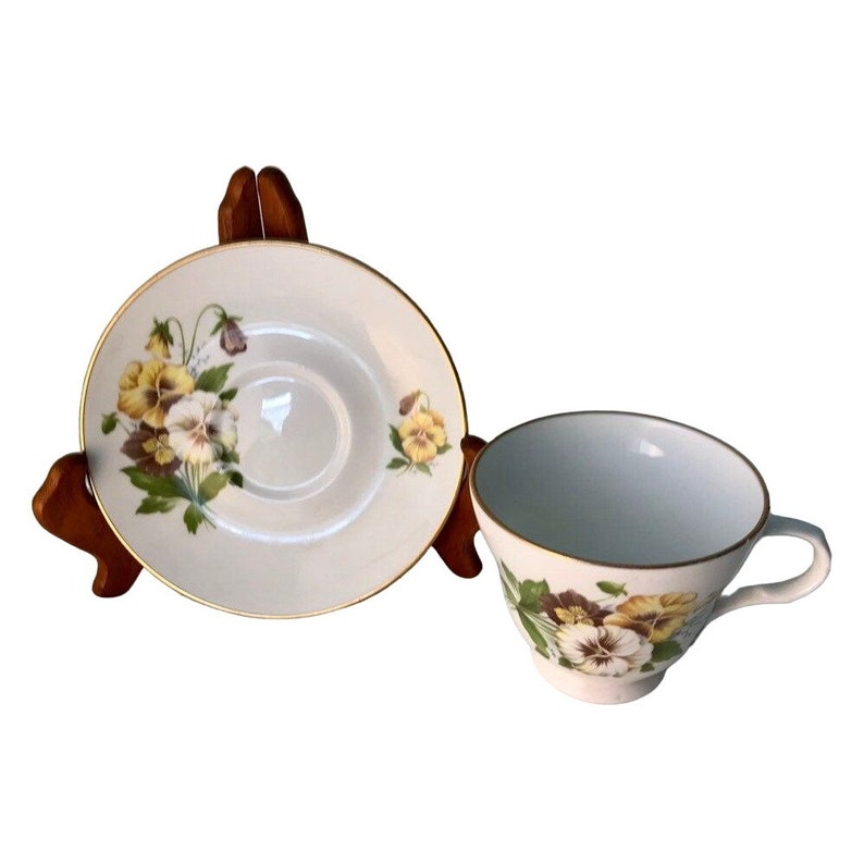May include: A white teacup and saucer set decorated with yellow and purple pansies. The cup and saucer have gold edging. The saucer is displayed on a wooden stand.