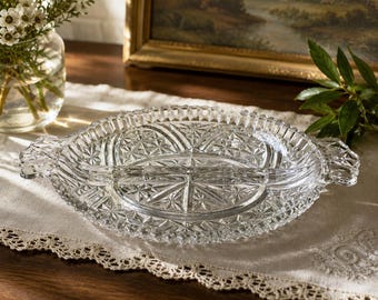 May include: A clear glass serving dish with a divided interior and two side handles. The dish features a pressed pattern of geometric stars and radiating lines, with a scalloped edge around the entire perimeter of the circular tray.