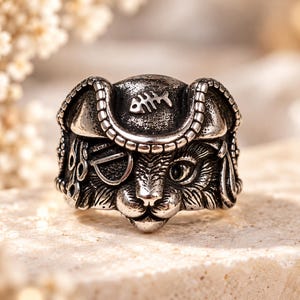 May include: A silver-toned metal ring featuring a detailed cat face wearing a pirate hat. The hat is adorned with a fish skeleton emblem, and the cat has an eye patch over one eye. The metal has a dark, oxidized finish to highlight the texture.