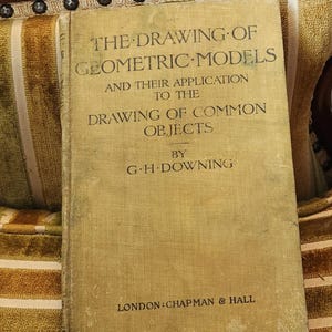 May include: A hardback book with a faded yellow-tan cover titled The Drawing of Geometric Models and Their Application to the Drawing of Common Objects by G.H. Downing, published in London by Chapman & Hall, featuring dark text on the front.