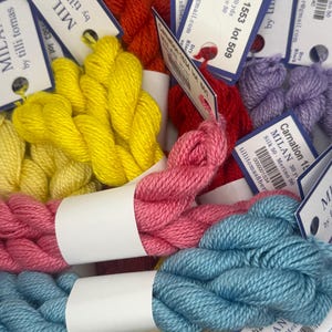 May include: A collection of Tilli Tomas Milan yarn skeins in various colors including yellow, pink, light blue, red, and purple. Each skein features a white paper label with black text detailing the color name, lot number, and fiber content.