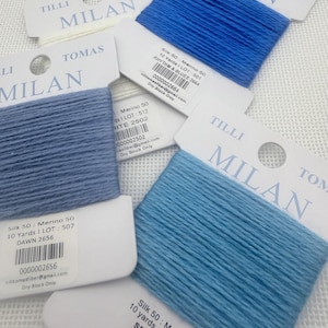 May include: Close-up of several spools of Tilli Tomas Milan silk yarn in various shades of blue and white. Each spool is labeled with the yarn type, color name, and lot number. The yarn is arranged on a white textured surface.