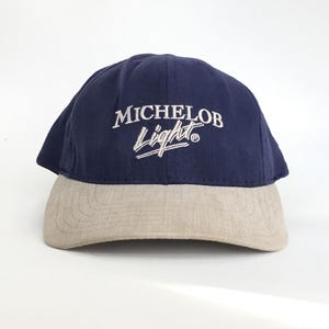 May include: A navy blue and tan baseball cap. The top of the cap is navy blue with the white embroidered text "MICHELOB Light". The bill of the cap is tan.