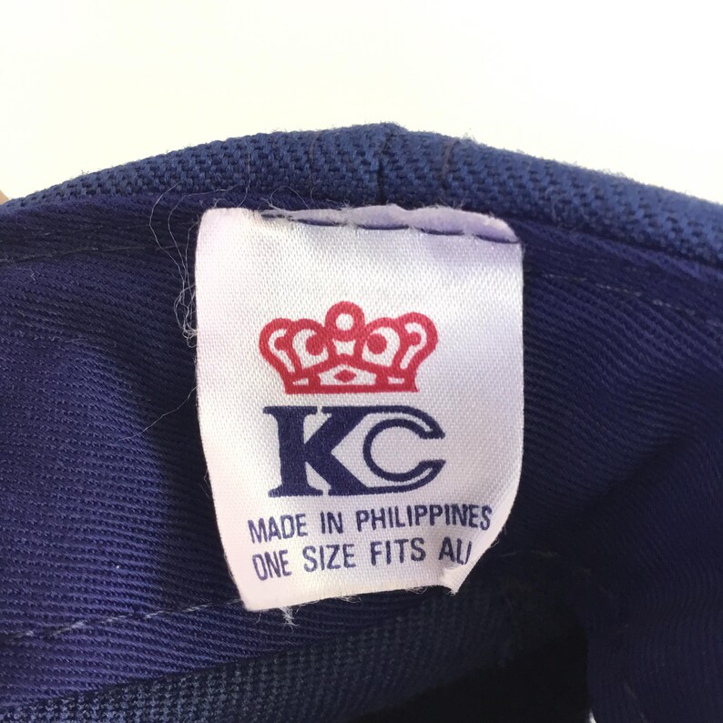 May include: Close-up of a blue baseball cap tag. The tag features a red crown above the letters "KC" in navy blue. Text on the tag reads "MADE IN PHILIPPINES" and "ONE SIZE FITS ALL".
