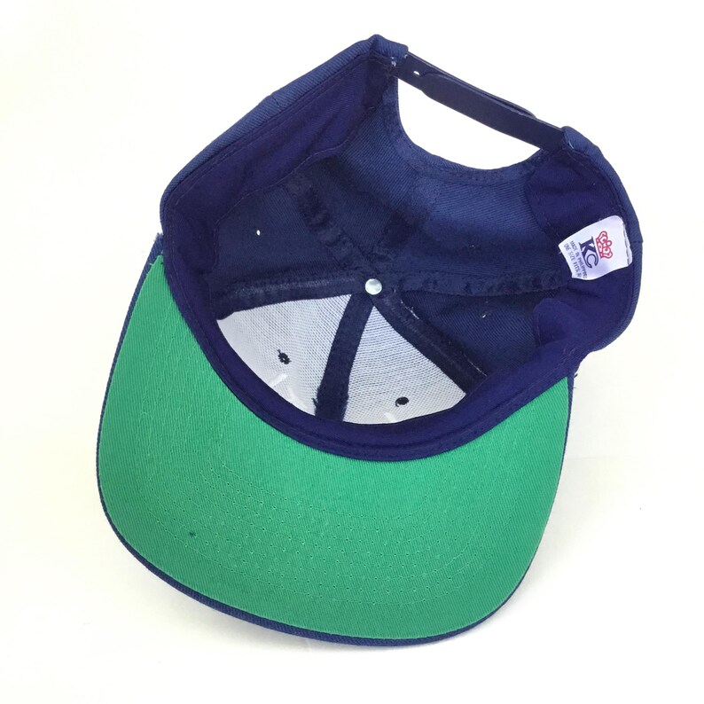 May include: A navy blue baseball cap with a green brim, viewed from the inside. The cap has a white mesh lining and a black adjustable strap. A small tag with the letters "KC" is visible.