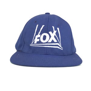 May include: A navy blue baseball cap with the word "FOX" in white block letters. The hat has a rounded crown and a curved brim. The hat is made of a durable fabric.