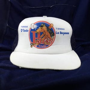 May include: White baseball cap with a curved brim and a mesh back. The front features a circular logo with a horse, baseball bat, and the text "Hot Gs" and "Tiendas D'todo La Sorpresa".