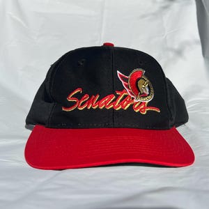 Puede incluir: Gorra de béisbol negra con visera roja. La gorra presenta la palabra "Senators" en escritura dorada, junto con un logotipo del equipo. La gorra está hecha de tela duradera.