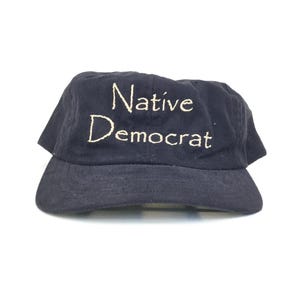 May include: A navy blue baseball cap with the words "Native Democrat" embroidered in a cream-colored thread. The cap has a curved brim and a classic six-panel design, suitable for casual wear.