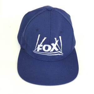 May include: A navy blue baseball cap with a curved brim. The cap features the white "FOX" logo on the front. The hat is made of a durable fabric and has a structured fit.