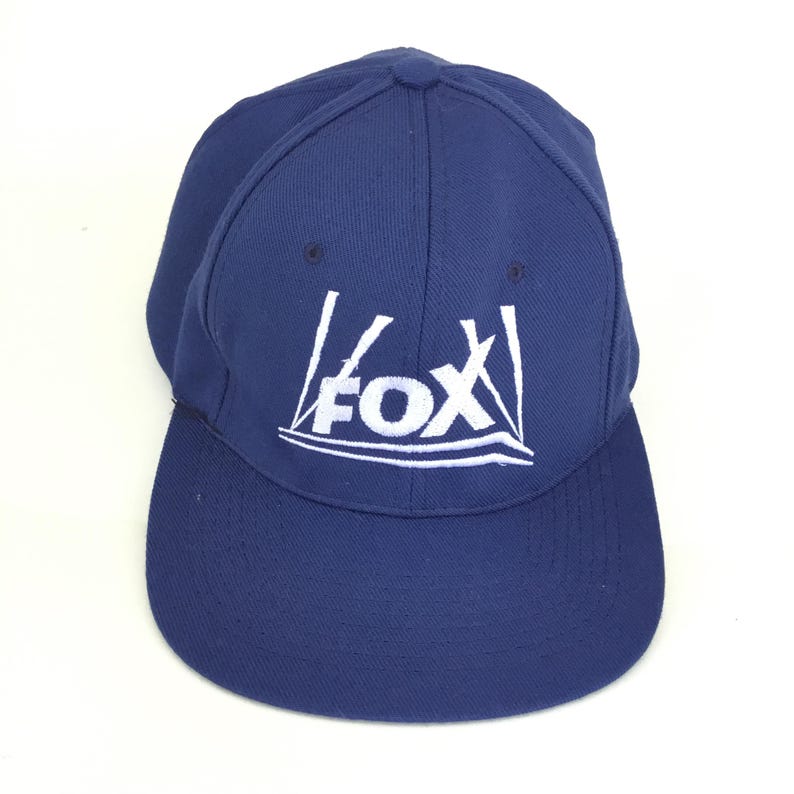 May include: A navy blue baseball cap with a curved brim. The cap features the white "FOX" logo on the front. The hat is made of a durable fabric and has a structured fit.