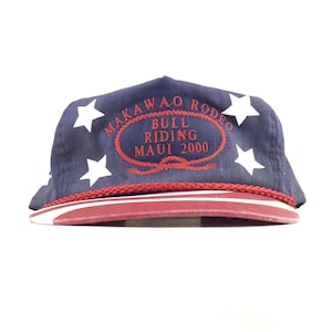 Könnte beinhalten: Eine marineblaue Baseballkappe mit weißen Sternen und roter Stickerei, die Makawao Rodeo Bull Riding Maui 2000 in einem seilartigen Oval zeigt. Die Kappe hat eine rote geflochtene Kordel an der Basis und einen roten Schirm mit weißen Streifen.