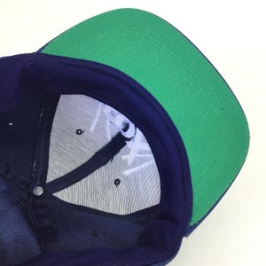 May include: A navy blue baseball cap with a green brim. The underside of the cap features white embroidered text and a mesh lining. The cap has a snap closure at the back.