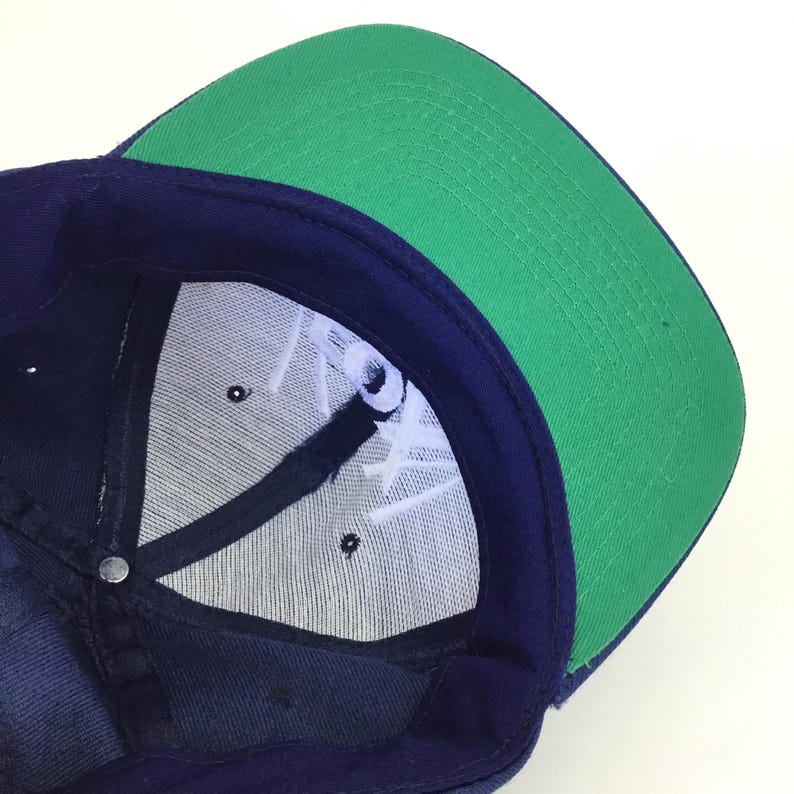 May include: A navy blue baseball cap with a green brim. The underside of the cap features white embroidered text and a mesh lining. The cap has a snap closure at the back.